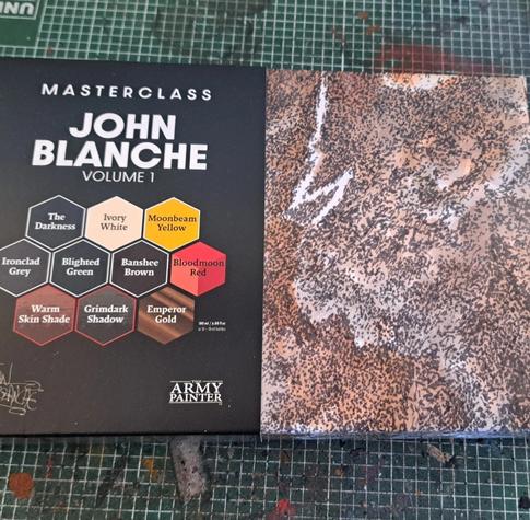 A box of paints. The text reads "Masterclass John Blanche Volume 1". There are 10 colours in hexagons ranging from dark bowns/greens to bright yellow