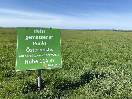 Weite Landschaft am Neusiedler See, im Vordergrund ein Schild mit Text:
tiefst gemessener Punkt Österreichs, am Schnittpunkt der Wege Höhe 114 m. APETLON, Lange Lacke