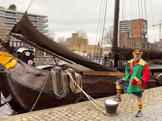 Bearded man, dressed up as a clown, but wearing running shoes and a kilt, in front of a wooden shop