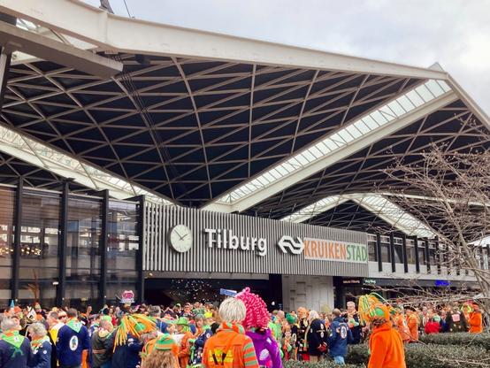 A view of the front of Tilburg train station, a colourful carnival crowd in front of it. Next to the official Tilburg NS sign, a banner with the city’s carnival name “Kruikenstad”