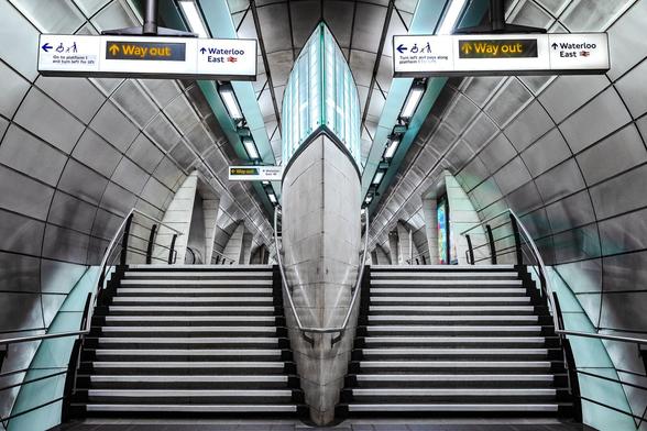 Symmetrical view of stairs leading to platform in circular corridor at Southwark Tube Station in London with an almond shape central pillar holding lit direction sides on each side. This perspective creates an illusory perception that the viewer is looking at something similar to an eagle face (pareidolia phenomenon). The icy blue lights on the top the central pillar and on the sides of the stairs accentuate that effect.