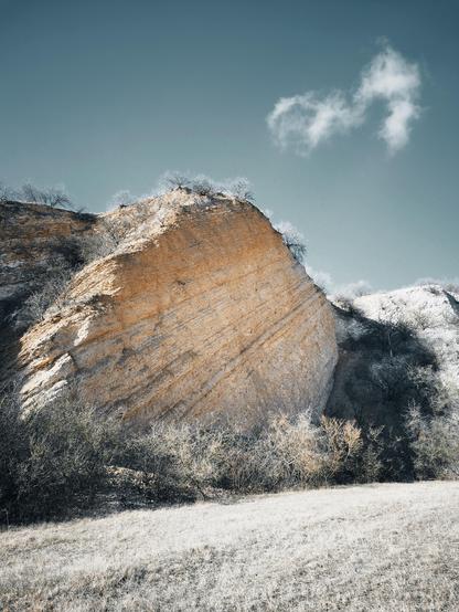 The image shows a large rock formation with an angled, rugged surface. It is surrounded by sparse vegetation, including bushes and small trees. The sky in the background is clear with a single fluffy cloud, giving the scene a peaceful and natural appearance.