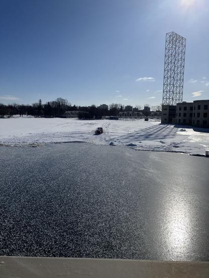 Blue sky overlooking a segment of river that has a square area obviously de-iced and refrozen with snow covered ice all around on three sides. there is a truck and trailer on the snow covered ice just ebyond the open water / refrozen segment. in the background sits a white bridge and a metal tower. The sun reflects on the ndewly frozen dark segment of the river.