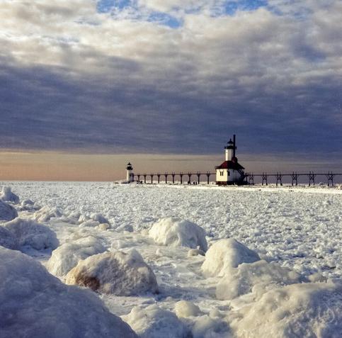 The setting sun lights up the frozen lake with the St. Joseph lighthouse in the background. The sky is dark and overcast.
