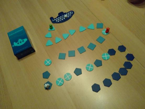 Auf einer Tischplatte links eine kleine Spieleschachtel in dunkelblau, mit Spieltitel und U-Boot-Illustration in hellblau. Rechts oben ein dunkelblaues Pappplättchen in U-Bootform, so groß wie die Schachtel. Darauf 25 kleine weiße Kreise, die über eine geschlängelte weiße Linie miteinander verbunden sind. Auf einem Feld ein weißer Holzstein. Vom U-Boot schlängeln sich viele eckige Wegeplättchen einen Weg nach unten, die in 4 Farbstufen immer dunkler werden. An manchen Stellen befinden sich runde Plättchen mit einem Kreuz. Drei Holzfiguren in Tauchyform stehen auf jeweils einem der Plättchen, davon eines in grün auf dem Plättchen direkt am U-Boot.