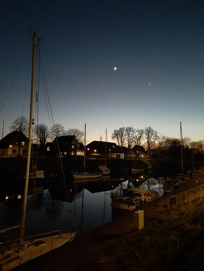 Beleuchtete Häuser und kahle Bäume vor Abendhimmel mit Sichelmond.
Im Vordergrund eine Anlegestelle an einem schmalen Fluss mit Segelboot und Motorbooten.