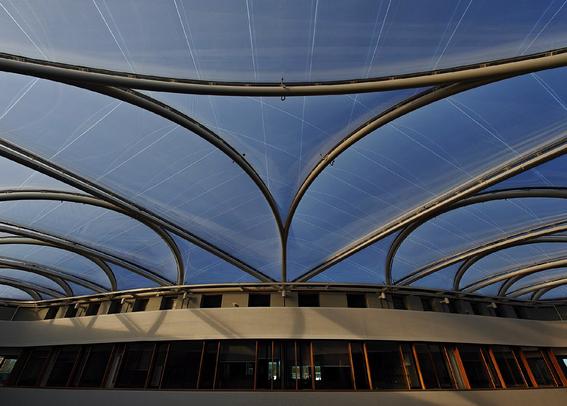 Modern architectural structure with curved metal framework supporting translucent roof panels, looking out onto a blue sky. The building has a beige inside facade with glass windows below.