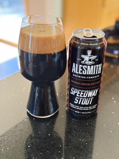 A stout glass full of dark beer with a thick tan head. The black can has brown highlights and white lettering with the AleSmith anvil logo top-center