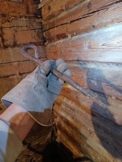 A gloved hand holding up a giant rusty spike that was hammered into a log beam wall that's visible behind. It has a square cut pointy tip and the rear is bent into a ring. It's about 30 cm long and perhaps 12mm diameter.