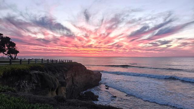 This photo captures a stunning sunset over the ocean, showcasing a dramatic sky and a scenic coastal view. The horizon line is positioned in the upper half of the image, with the ocean stretching towards it, displaying gentle waves rolling towards the shore. 

The sky is the focal point, dominated by a vibrant sunset. Wispy, striated clouds fan out from the center, radiating in shades of pink, purple, and pale orange against a backdrop of a soft, gray-blue twilight. The colors are intense yet soft, creating a captivating display of natural beauty.

To the left, a dark silhouette of a tree stands against the colorful sky, its branches reaching upwards. Below it, a grassy cliffside descends towards the ocean. A wooden fence runs along the edge of the cliff, suggesting a walking path or viewing area. The cliff itself is rocky and rugged, leading down to a small strip of beach where the waves break. 

The ocean occupies the lower right portion of the image, its surface reflecting the colors of the sky. The waves are relatively small, creating a sense of tranquility and peace. The overall composition is balanced, with the land on the left and the ocean on the right, framing the breathtaking sunset in the center. The image evokes a sense of calm and wonder, highlighting the beauty of nature at dusk.