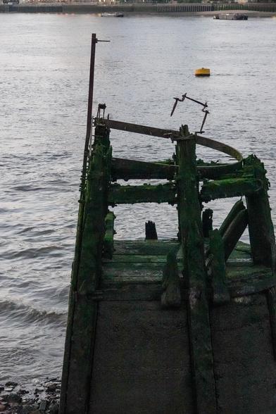 A wooden jetty in a sits on a stone base in a river, seen at night its beams are green and much damaged. There's a metal ladder attached to it that sticks out higher, and several metal objects attached at various angles.