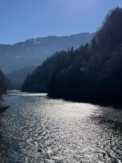 Ein ruhiger Fluss fließt durch eine bergige Landschaft unter einem klaren blauen Himmel. Sonnenlicht scheint auf die Wasseroberfläche und erzeugt einen schimmernden Effekt, während Bäume die Flussufer säumen und sich im Hintergrund Berge erheben, teilweise mit Schnee bedeckt.