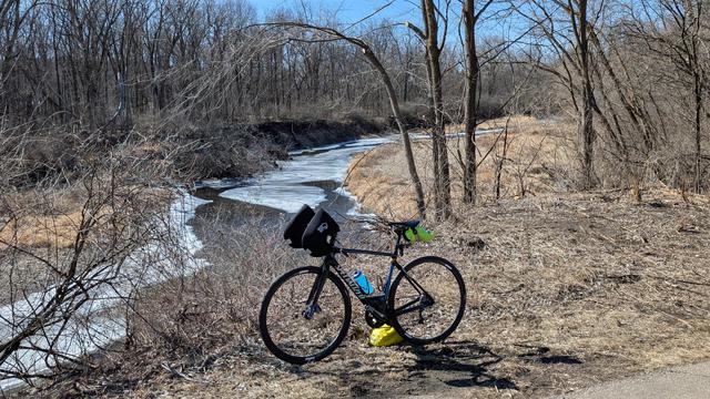 Blue and black Roubaix bicycle parked off the Zumbro River South Trail with the partially frozen river behind with brown lawn and bare trees surrounding. The sky is blue.