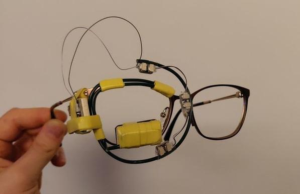 Ein Eye-Tracking-Brillengestell in Nahaufnahme. Es handelt sich um ein Brillengestell mit Ärmchen aus Gartendraht, Minikamera, und kleinen Infrarot-LEDs. Klebeband und Kabel lassen das Gestell wie einen Prototypen aussehen.