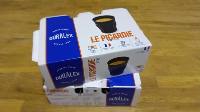 Photographie de deux boites de six verres noirs de 9 cl sur une table en bois. Le packaging des deux boites est en carton. On peut y voir sur le côté droit une bande bleue ou est inscrit le logo de la marque en blanc. Une image du dit-verre rempli de café (on peut reconnaître sa mousse). Dessous il est écrit le nom du modèle, "Le Picardie", en couleur orange et "Expresso", en bleu (le même que la bande de gauche). Dessous et enfin, il y a 4 dessins et informations : "Résistant, verre trempé" avec deux verres dessinés qui s'entre-choquent mais ne se brisent pas. Trois bandes cote-à-cote (bleue, blanche, rouge)  et "Fabriqué en France". Le dessin d'un verre grisé, en-dessous duquel il est inscrit "Verre teinté masse". Puis un dessin de verre avec une fumée colorée du même orange que "Le Picardie". En dessous de ce dessin, il est inscrit "Idéal boissons chaudes". Ces annotations sont également  traduites en anglais en dernières lignes et d'une police plus petite.