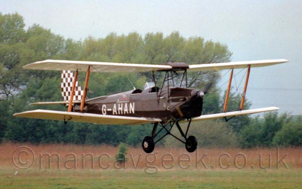 Side view of a brown, single-engined biplane flying from left to right at a very low altitude.
The plane has a pair of open cockpits in the top of the fuselage, both of which appear to be empty, at least at first glance, there is a hint of someone sitting in the rear cockpit.
The registration "G-AHAN" is on the rear fuselage in white, while the tail is covered in a brown and white checkerboard pattern.
Green grass fills the bottom of the frame, with trees in the background, under grey skies.