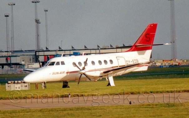 Side view of a white, twin propellor-engined airliner taxiing from right to left.
The plane has a gold and red stripe running along the body, overlaid with black "Sun Air" titles on the forward fuselage under the cockpit windows.
The stripe is repeated on the sides of the engine cowlings.
The tail is red, with a black letter "S" in the middle.
The registration "OY-EDB" is on the side of the rear fuselage in black.
Green grass fills the foreground, either side of a taxiway, with a grey terminal building stretching across most of the frame from the left.
Tall, skeletal lighting towers reach into the sky, with grey clouds filling the rest of the frame.