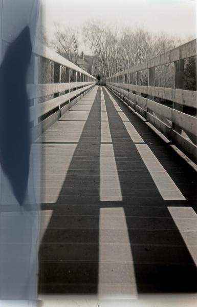 A grainy black and white image, looking down a long wooden bridge. The sun has cast dark shadows on the walkway. A large smudge is visible down the left side of the picture.