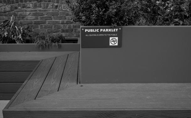 A black-and-white image showing part of a bench which bears a plaque reading "PUBLIC PARKLET / all seating is open to the public" with a little no-smoking icon.  There are plants and a brick wall in the background.  The white lettering on the dark plaque pops visually.
