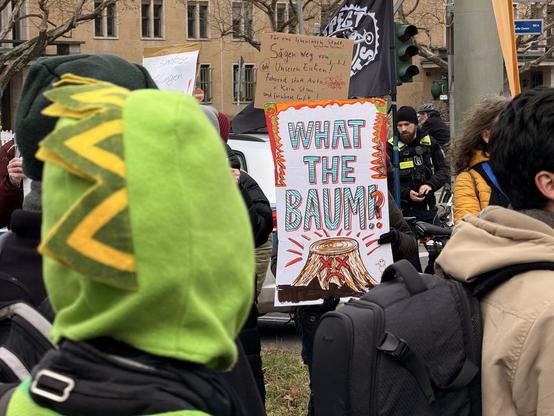 Foto eines Demo-Schildd auf dem ein Baumstumpf mit x-Augen zu sehen ist und Text „What the Baum!?“. Im Vordergrund ist ein Mensch in einem Drachenkostüm von hinten zu sehen.