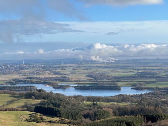 View over Loch Ore with forests in the foreground, wind turbines and a town in the background.