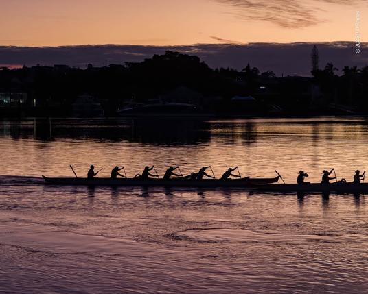 Looking across the Brisbane river at dawn, a coppery sky reflecting darker in the lightly rippled surface. Two six-person canoes are paddling up-river, silhouetted against the bright water, bodies, arms and paddles all synchronised. The opposite bank of the river is dark, just a few lights visible, and only the outlines of tree and rooftops apparent.