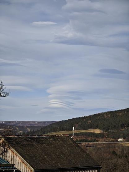 Picture taken down the valley, garage roof in foreground. Above the junction of two mountains to the centre, a weird stack of clouds has formed. Three very smooth short ovals packed close together vertically. Possibly the pattern is repeating in some additional larger clouds above.