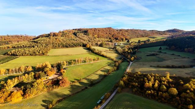Drone picture taken at the Gierskopp in autumn showing the view to the Restaurant Schinkenwirt with lots of cloured trees and a blue sky with some clouds in the background