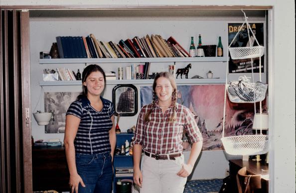 Two college aged women pose together in a sleeping area that's accessible by an accordion door. The woman on the left has dark hair and is wearing a dark blue and gray short sleeve shirt and blue jeans. The woman on the right has auburn hair and is wearing a long sleeve red plaid shirt with the sleeves rolled up past her elbows, and light colored trousers. Behind them are shelves and movie posters.