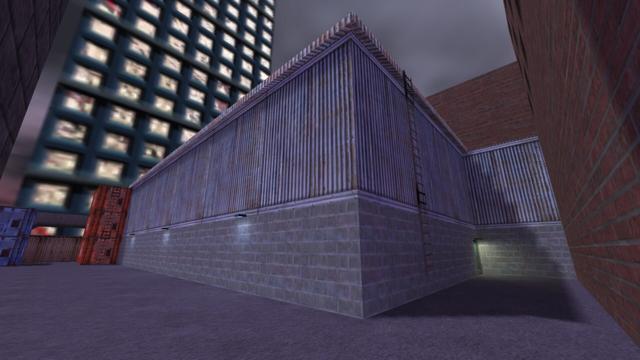 In this picture you can see the previously mentioned back entrance into the warehouse. Above this is an yellow illuminated lamp. Next to the back enterance is a ladder. To the left of the long side of the warehouse, are three white illuminated lamps. Furthermore, on the left you can see three stacked red containers, on which there is a ladder, and two blue stacked containers. Behind it, in the skybox, you can see a skyscraper. This is on the map cs_assault in the game Counter-Strike 1.6.