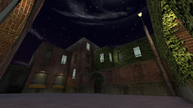 In this picture you can see a red brick building with two loading zones for trucks. Above each of these is a yellow illuminated lamp. All windows of the building are provided with bars. In some rooms lights are on. Except where instead of two windows there are two wooden boards. There are also no bars in front of them. On the right side of the building, ivy grows all over the wall. To the left of the building you can see a green garbage container. Furthermore, you can see two entrances on the left and right edge of the picture. In addition, in front of the part of the building with ivy there is a wall with a passage. In front of the wall is an yellow illuminated lamp attached to a wooden pole. This is on the map cs_backalley in the game Counter-Strike 1.6.