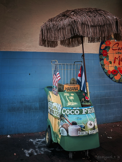 Little cart that sells fresh coconuts in front of the wall painted on white and blue. There’s a straw parasol attached the cart. US and Cuban flags are hanging on both side of the basket on top of the cart