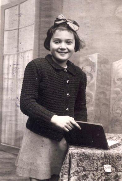 Black and white photo of a girl standing beside a table, smiling at the camera with a book in hand.