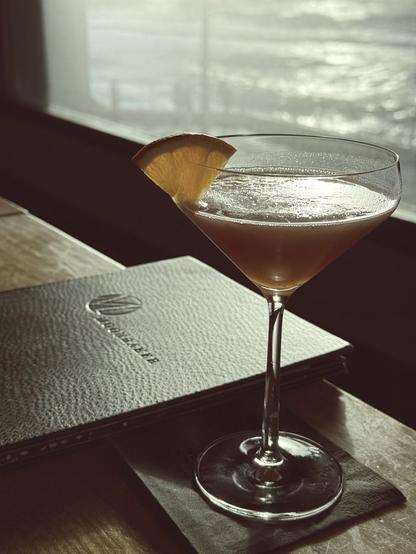 Cocktail in front of a menu on table with sunset reflected off the ocean outside window in the background