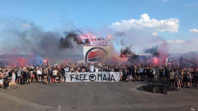 Hunderte Antifaschist*innen hinter einem Banner auf welchem in schwarzen Großbuchstaben "Free Maja" steht in der Mitte zwei schwarze Fahnen in einem Kreis. 

etwas im Hintergrund ein mehrere Meter hohes Podest auf welchem ebenfalls mehrere Antifaschisti mit Pyro stehen.