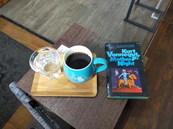 A bird's eye view of a small square wooden table on which the small black paperback book with "Kurt Vonnegut Jr " turquoise complimenting the turquoise coffee mug of black coffee with the white cat barista on the mug. The book has an illustration of an all turquoise long sleeve shirt & pants on a large brown dog. He is holding a red megaphone & has a white cowboy hat on and holds a nazi flag. The mug is on a small tray with a clear tumbler to the left.