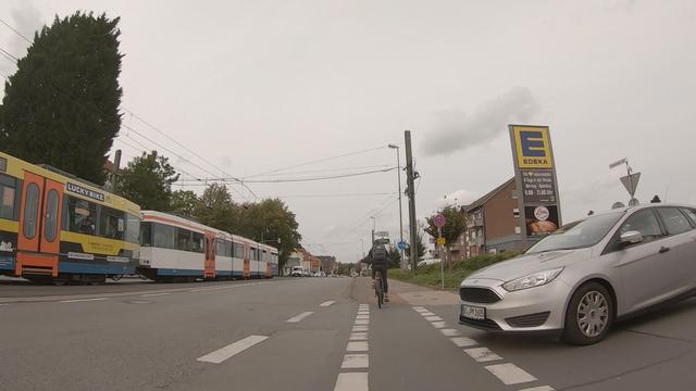 Dashcam-Aufnahme vom Rad aus. Straßenbahn links, daneben ein breiter Asphaltfahrstreifen, dann sehr schmal benutzungspflichtiger Radweg und Fußweg. von Rechts will ein Auto vom Edekaparkplatz runter.