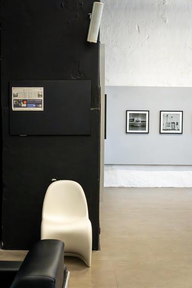 Blick auf eine Ecke in den Räumen der GAF. Ein weißer Stuhl vor einer schwarzen Wand. Rechts davon ist der Ausstellungsraum samt Fotografien