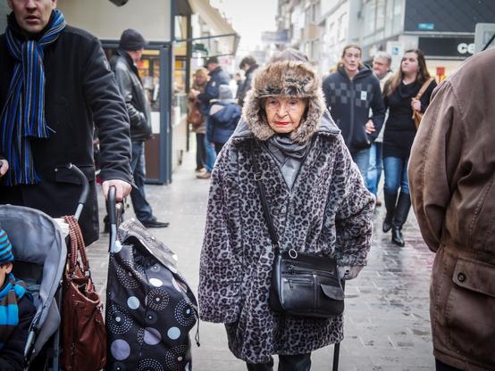 An eye-level medium shot shows an elderly woman in a fur hat and leopard print coat walking among a crowd of people on a city street. The woman is in the center of the shot, walking towards the camera. She is wearing a large fur hat, a gray scarf, and a leopard print coat. She is also carrying a black purse and holding a cane in her right hand. The people around her are dressed in winter clothing, including coats, hats, and scarves. To the left of the woman a man is pushing a stroller. The street is wet, and there are buildings on both sides. The sky is overcast.