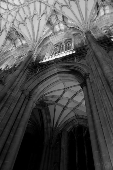 A black and white photo looking up at a cathedral's interior.  Thick, round columns line the right side, leading to a tall arched doorway with detailed stonework. Above the archway, a large window with Gothic tracery is visible. The vaulted ceiling dominates the image with its intricate fan vaulting, creating a complex pattern of ribs and arches. Light and shadow play across the stone, highlighting the texture and craftsmanship. The image conveys a sense of grandeur and the soaring height of the space.