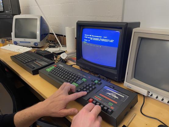 Langer Tisch mit drei alten Monitoren, einer Tastatur und zwei alten Mikrocomputern: ein iMac aus den 2000er Jahren, eine Sinclair Spectrum 2, und ein Amstrad CPC 464. Zwei Hände versuchen sich in einer Tastenkombi auf dem Amstrad.