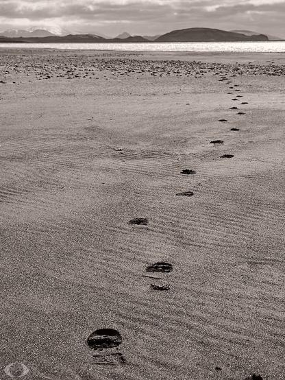 A black-sand beach on an ice lake in Iceland. Footsteps in the sand apparently going nowhere. Behind the lake snow covered mountains.