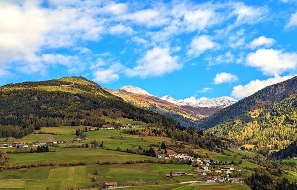 A scenic landscape photograph of a valley nestled between rolling hills or mountains. The valley floor is dotted with small houses, suggesting a village. The hills are covered in trees, with some patches of green fields and some areas where the trees are showing signs of autumn colors. In the background, there are higher mountains with snow-capped peaks. The sky is blue with scattered white clouds.