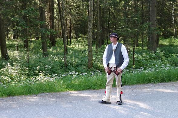 A Górale in traditional costume waiting to take tourists to Morskie Oko, Tatra mountains, in a horse cart. He is standing in the road that leads up to the lake and mountains. Behind him is pine forest. This costume is everyday wear.