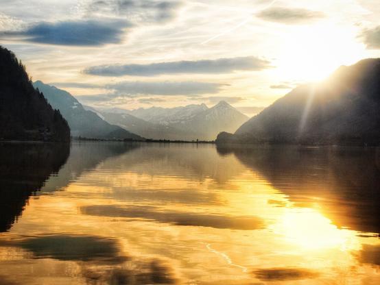This image captures a breathtaking sunset over Lake Brienz (Brienzersee) as seen from Iseltwald, Switzerland. The golden light of the setting sun reflects on the calm waters, creating a mesmerizing mirror effect that enhances the beauty of the surrounding landscape.

Framed by towering mountains, the lake stretches into the distance, with the snow-capped peaks adding contrast to the warm tones of the sunset. The sky displays a mix of soft clouds and clear patches, through which the sun’s rays filter, casting a dramatic glow over the scene. The composition balances the dark silhouettes of the hills with the luminous reflection on the water, highlighting the serene and picturesque nature of this iconic Swiss location.