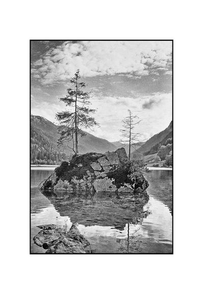This black and white portrait photograph shows two trees growing on a rock protruding from the water.
In the foreground on the left are stones protruding from the water. Behind them, the large rock and a tree are reflected in the water.
The large rock is in the center of the picture, slightly below the horizontal center of the picture. It is completely surrounded by water.
Two conifers grow on the barren rock. One on the left side of the rock, the other on the right.
There is a mountain landscape in the background. Tree-covered mountains rise steeply to the left and right of the rock. Behind the rock is a valley.
Above the mountains and the rock, clouds and a clear sky divide the background.