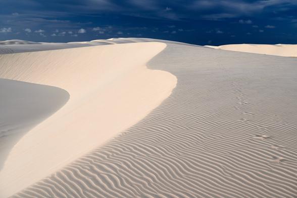 A sand dune is shown with the ridge of the dune leading from the left down corner into the upper middle of the photo. In the background further dunes are visible. Above the dunes is a dark blue sky with patches of white clouds.
