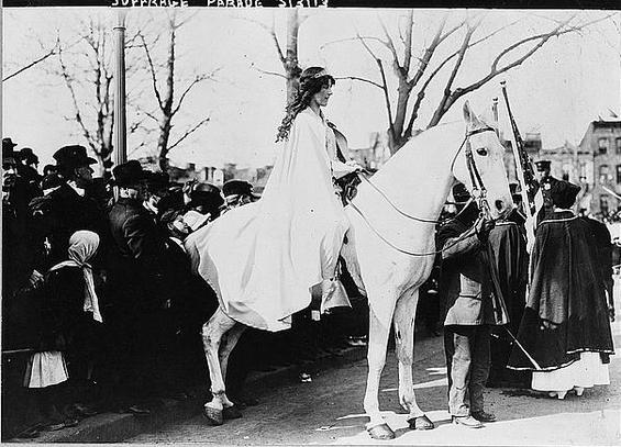 A white woman on a white horse, wearing a white robe. The horse's reins are being held by a man, and a crowd stands behind them.