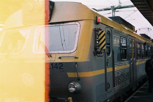 A grainy analog photograph of an electric train locomotive sitting at a train station. The train is a model X11 from 1985, painted in Swedish train operator Västtågen's grey and yellow livery. It has a combination of boxy and rounded features, including some classic, round headlamps. The left third of the picture is covered in a bright yellow light leak, showing that this was the first photo on this roll of film.