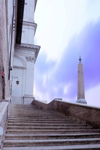 Eine Treppe führt aus dem Vordergrund entlang einem historischen Gebäude hinauf. Im Hintergrund ist ein Obelisk und ein blau lila himmel sichtbar. / A staircase leads up from the foreground along a historic building. An obelisk and a blue and purple sky are visible in the background.